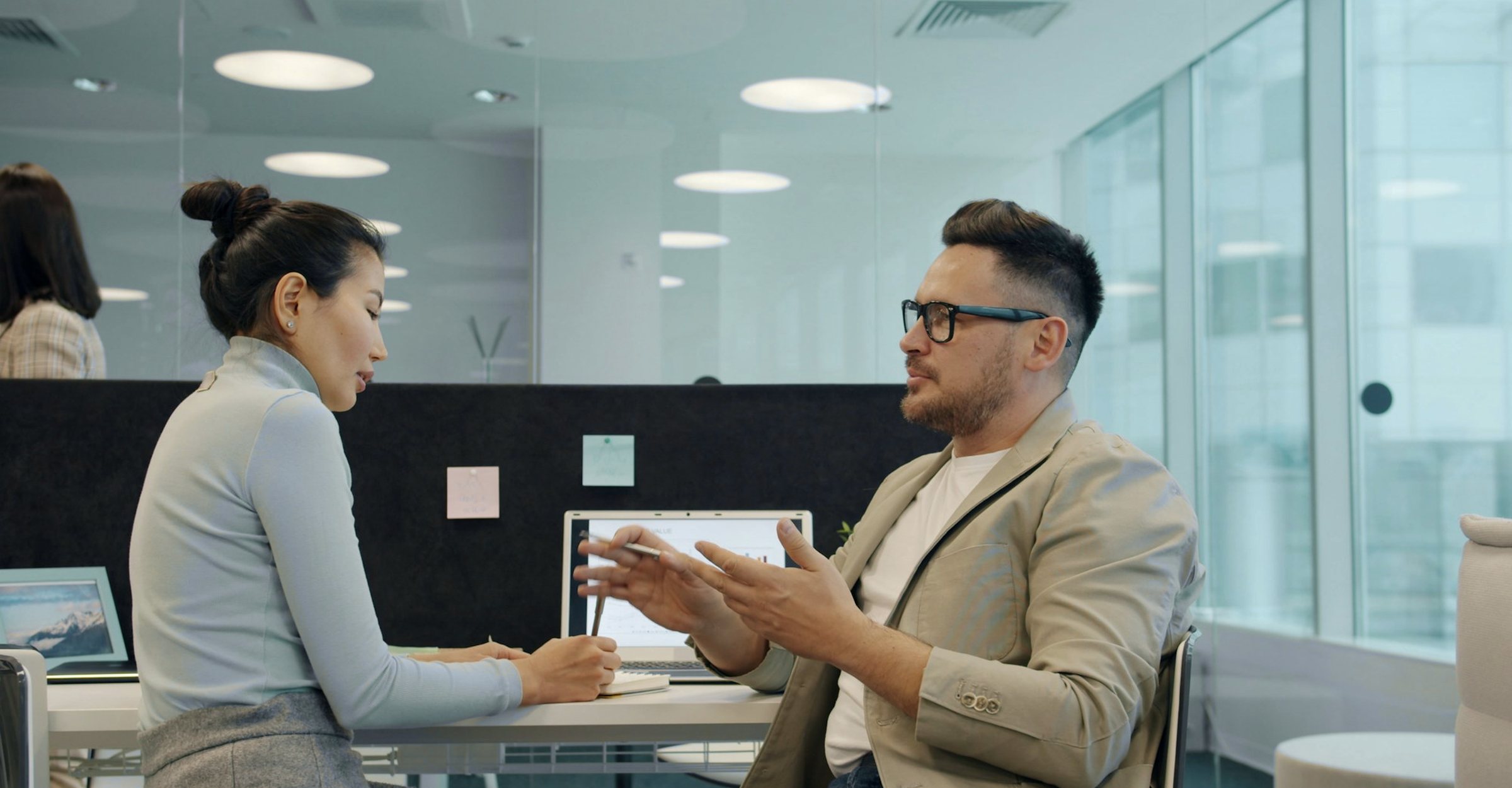 Two colleagues talking at a desk in a glass-walled office